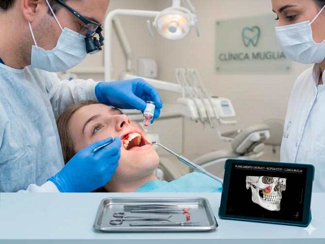 Dentista em procedimento de cirurgia para biópsia e remoção de cisto na Clínica Muglia, com planejamento digital em tablet.
