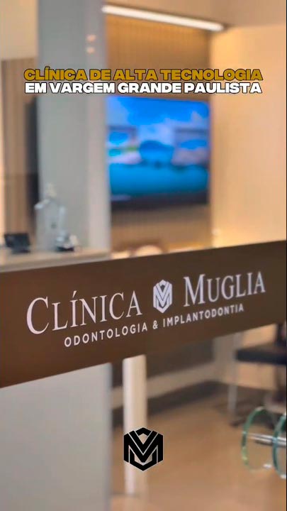 Fachada da Clínica Muglia Odontologia com porta de vidro, faixa marrom com logotipo branco, recepção ao fundo com iluminação amarela aconchegante e tela digital exibindo paisagem de céu azul, clínica odontológica em Vargem Grande Paulista